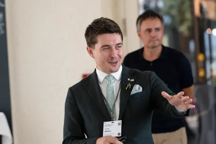 A man in a suit gestures while speaking, with another man in the background.